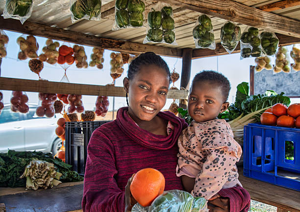 Happy African street vendor holding her child, shack with vegetables and fruits, women empowerment with skills Private Sector Engagement direct from farm sale